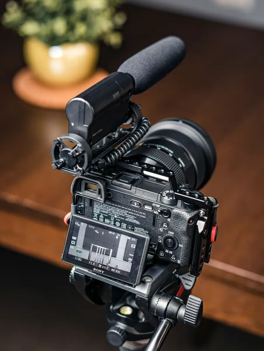 Caméra Sony avec microphone monté et écran LCD affichant les paramètres d'enregistrement, posée sur un trépied en bois.